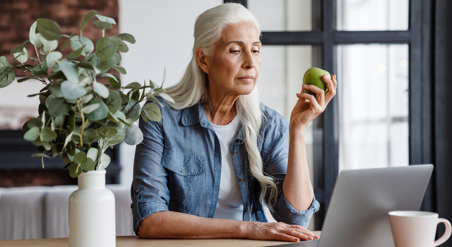 senior woman with long gray hair and blue shirt eating green apple in front of laptop computer
